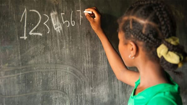 A young girl facing a chalkboard writing her numbers on the board from 1 to 7