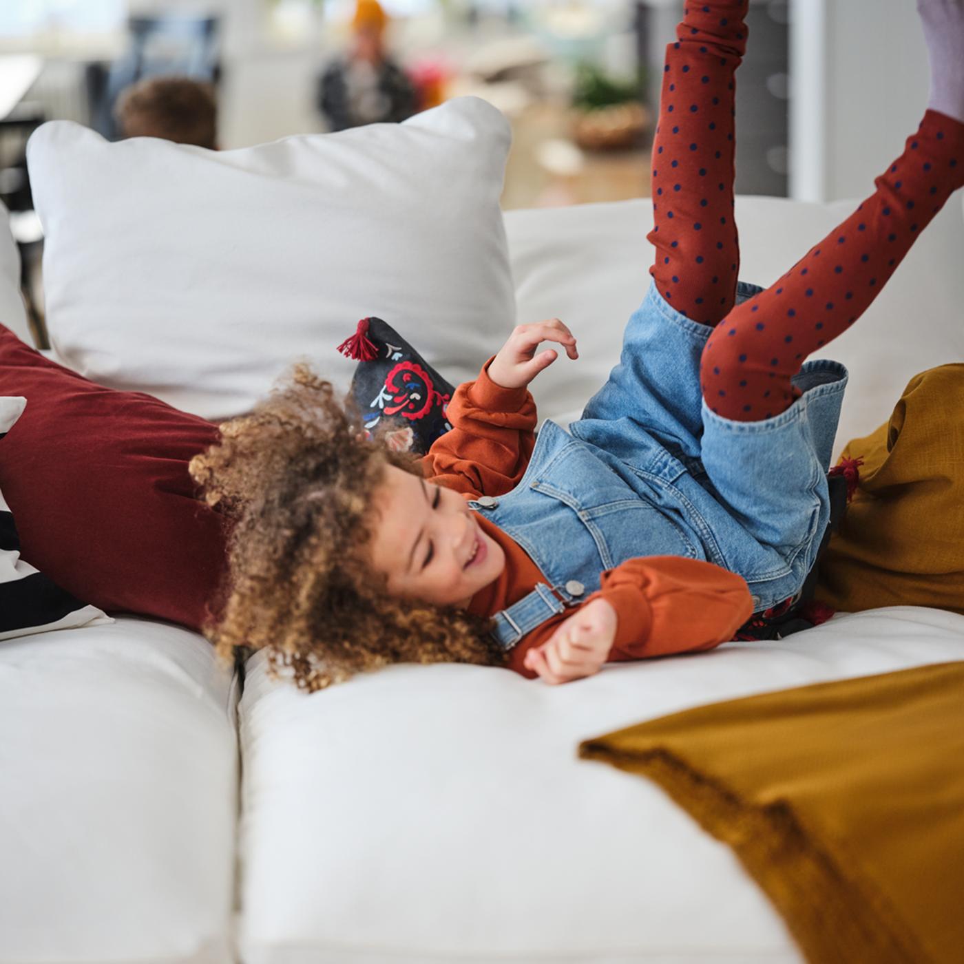 A young girl rolling around, feet in the air, amid colourful textiles on a white GRÖNLID four-seat sofa with chaise longues.