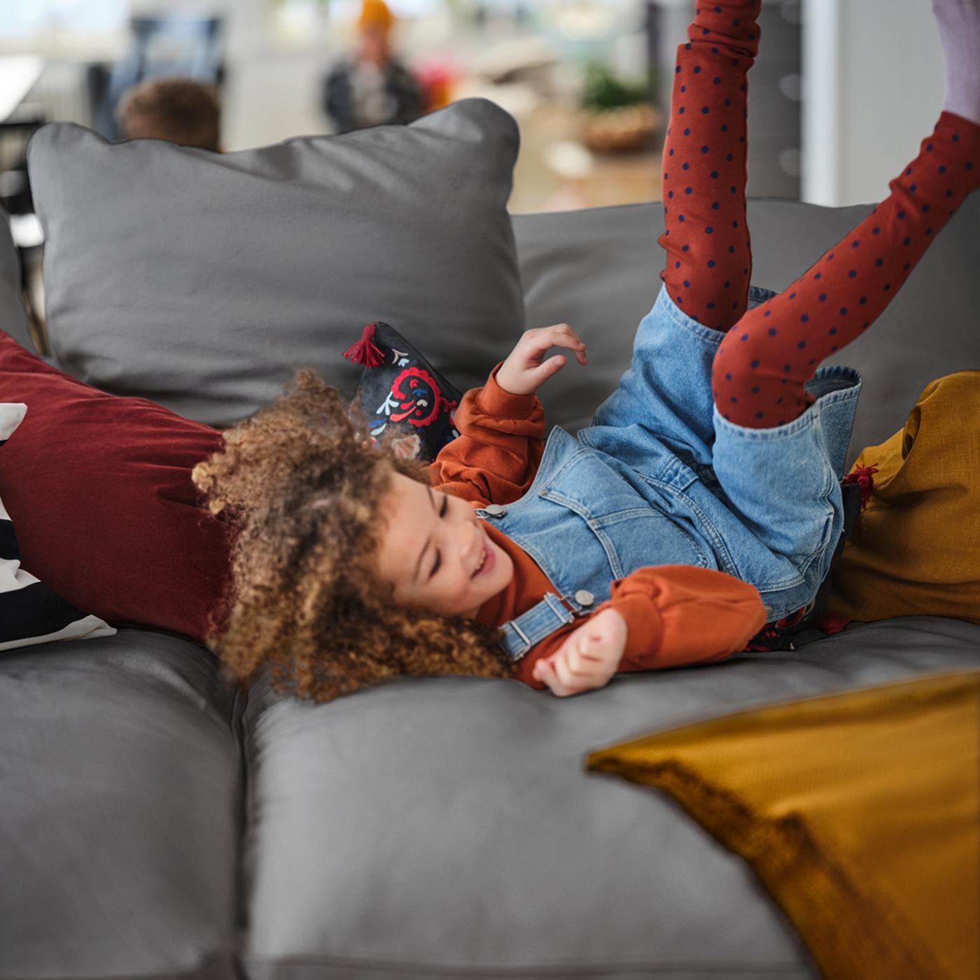 A young girl rolling around, feet in the air, amid colourful textiles on a grey GRÖNLID four-seat sofa with chaise longues.
