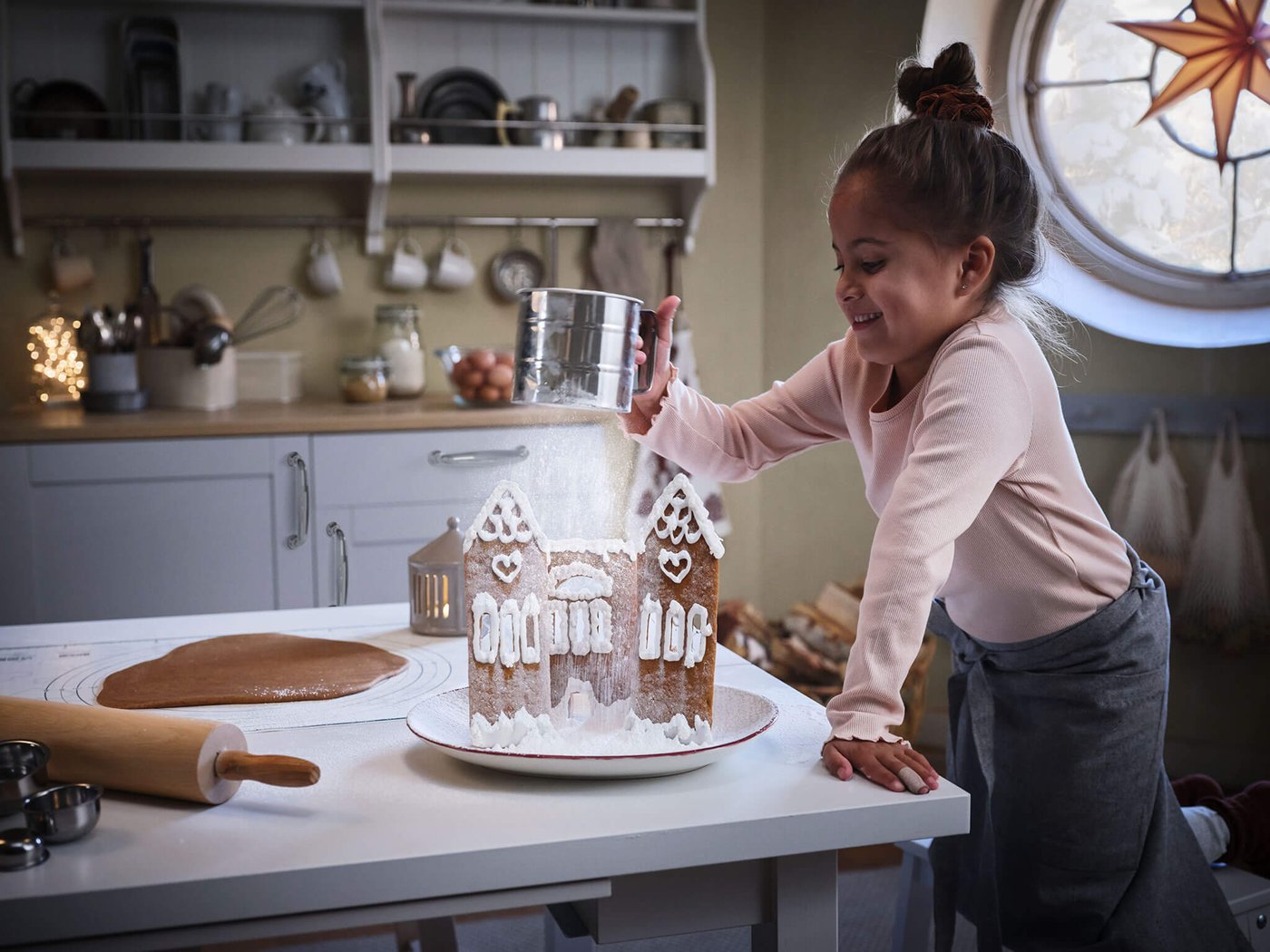 A young girl sifting powdered sugar on a gingerbread house in a kitchen.