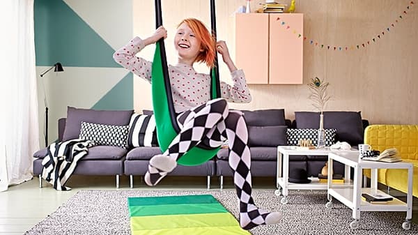 A young girl sitting in a swing, in the living room, with a gym mat underneath.