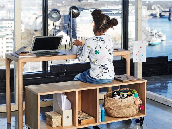 A young girl sitting on a wooden bench at a desk with a laptop and lamp in front of a large window.