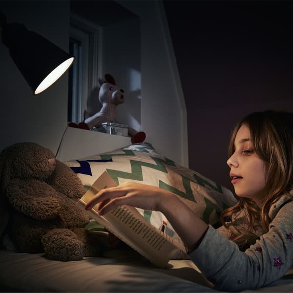 A young girl, wearing a grey sweater, lying in bed and reading a book under a bright light.