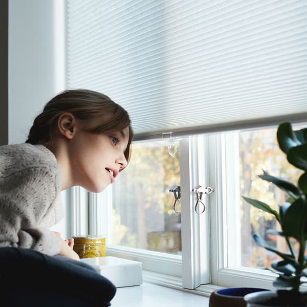 A young girl, wearing a sweater, looks out of a window underneath a partially pulled down blind.