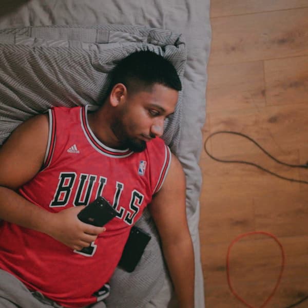 A young man with a beard and a red sports shirt is relaxing on a grey sofa while holding a phone.