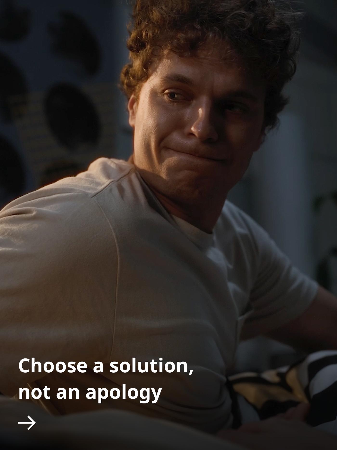 A young man with curly hair sits indoors in warm lighting, looking emotional but composed, with text reading “Choose a solution, not an apology.”