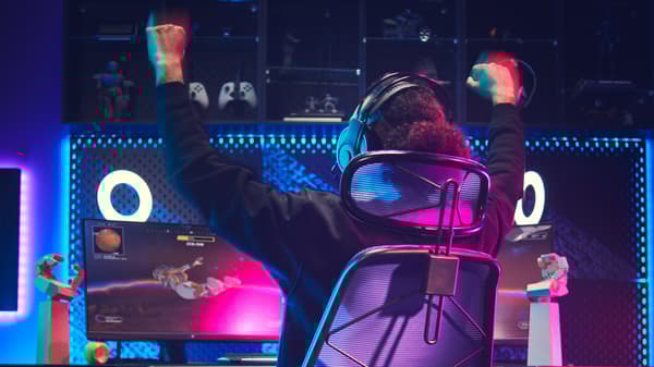 A young man with headphones on and his hands up in the air sits in a STYRSPEL gaming chair in front of two screens.