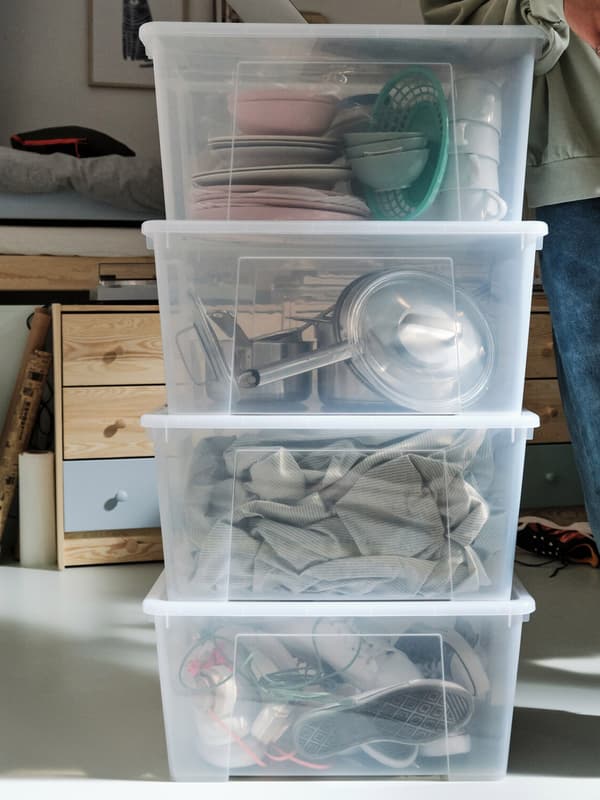 A young person next to four transparent 45 l SAMLA boxes stacked on top of each other, filled with shoes and cookware.