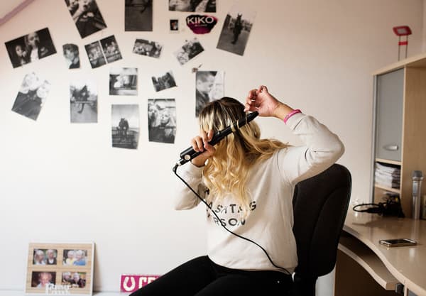 A young woman curling her hair with an electrical curler in her white-walled bedroom.