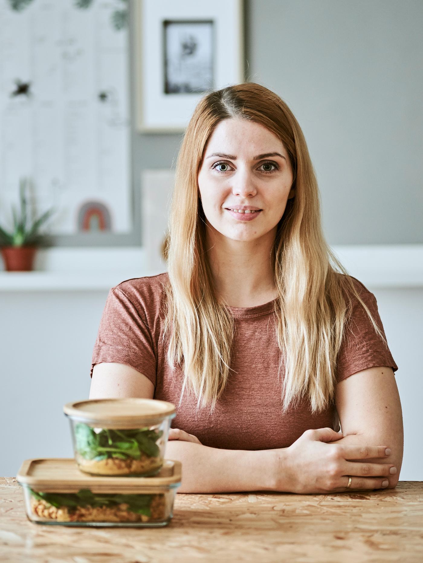 A young woman sits at a wood table with two bamboo-lidded glass food containers filled with lentil stew and spinach.