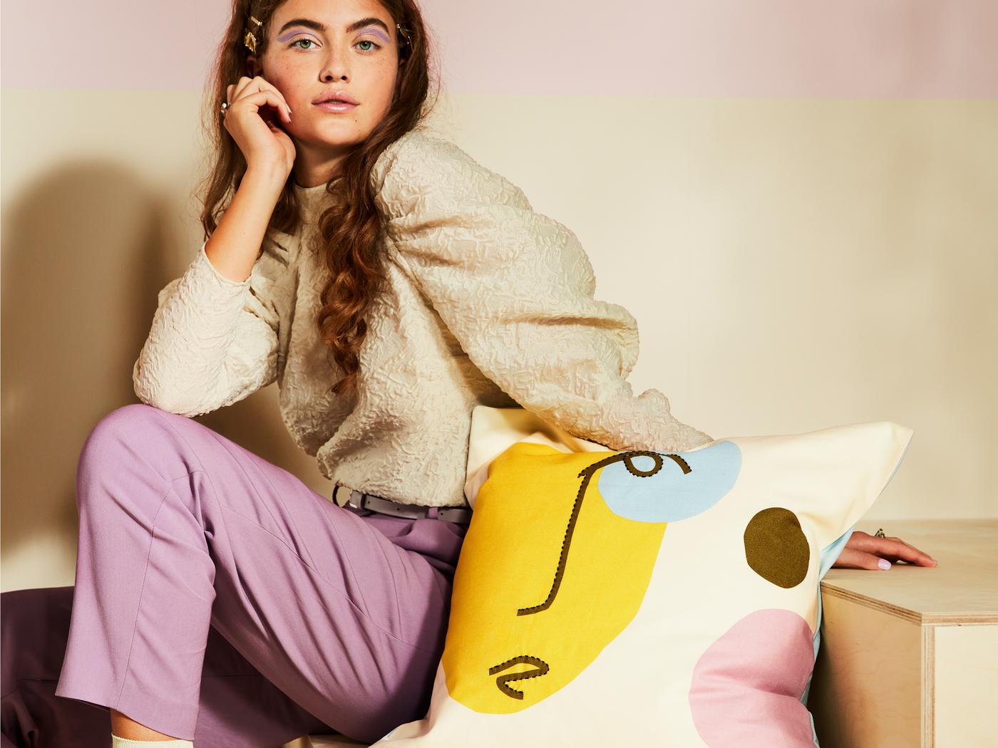 A young woman sitting on a wooden staircase with a cushion featuring an abstract pattern of a face with bright colours.