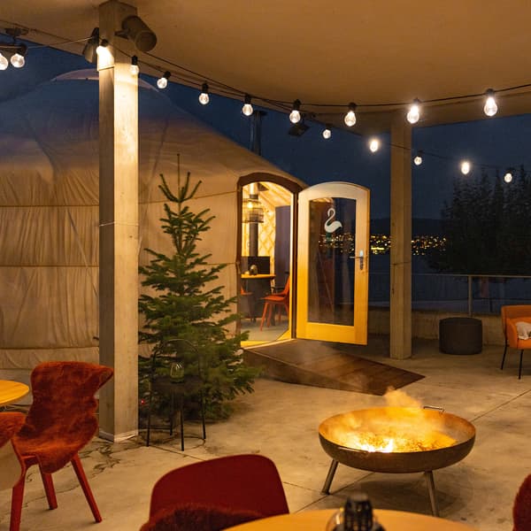 A yurt with an open door in front of a terrace with chairs, tables and a fire bowl