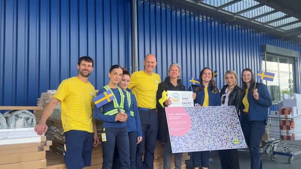 Acht Personen in gelb-blauer Kleidung stehen vor einem IKEA Einrichtungshaus, halten kleine schwedische Flaggen und ein großes Plakat mit Text und Bildern einer Spende. Um sie herum befinden sich Kartons und Gegenstände.
