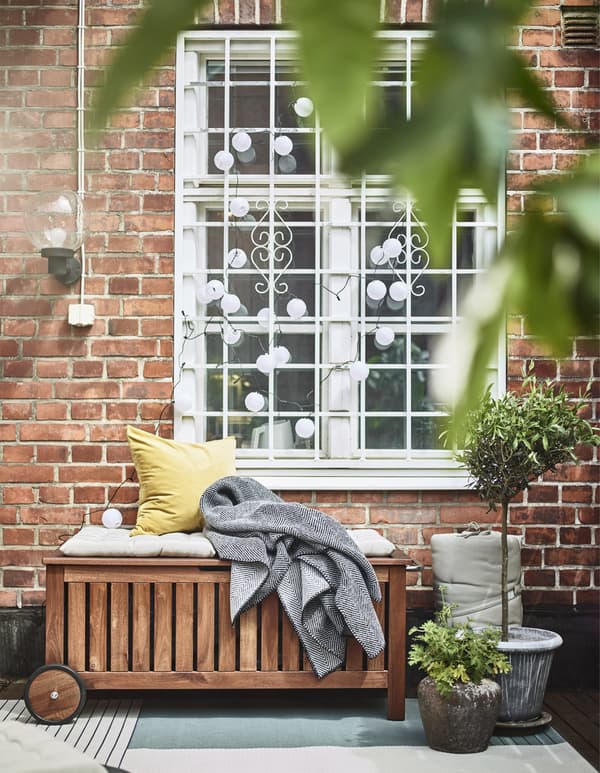 ÄPPLARÖ/TOSTERÖ bench with IKEA storage bag positioned outside a window, shown with cushions, a throw and potted plants.