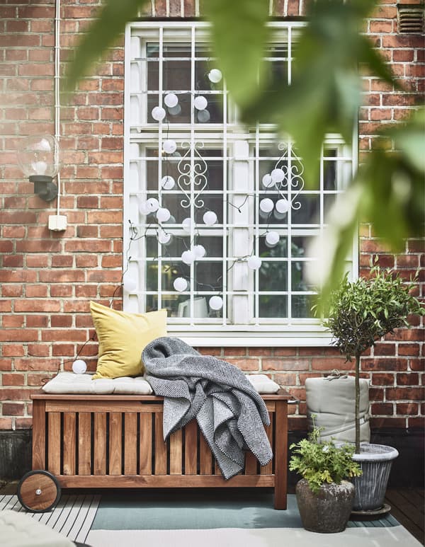 ÄPPLARÖ/TOSTERÖ bench with storage against a brick wall with hanging white lantern lights and textiles ontop.