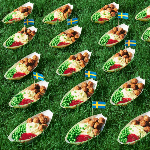 Against a background of green grass there are several long dishes filled with meatballs, mashed potatoes, and green peas. Swedish flags are attached to the dishes.