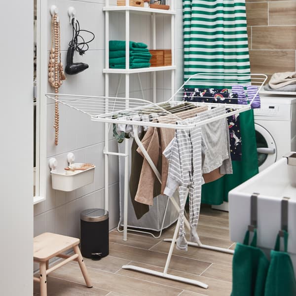 علاقة تجفيف MULIG بيضاء مع جناحين يفتحان للخارج أمام غسالة وملابس معلقة لتجف.