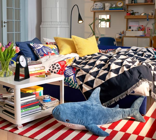 Alas tenunan jacquard JOHANNE dengan corak hiasan di atas katil sofa di samping lampu dan meja sisi katil yang mempunyai banyak buku di atasnya.