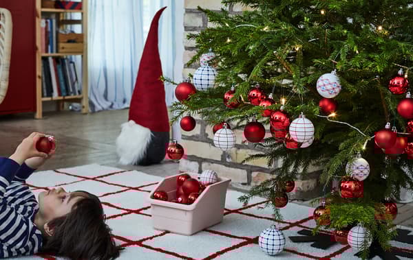 Albero di Natale pieno di decorazioni e un bambino sdraiato a pancia in su sul tappeto mentre osserva una decorazione natalizia - IKEA
