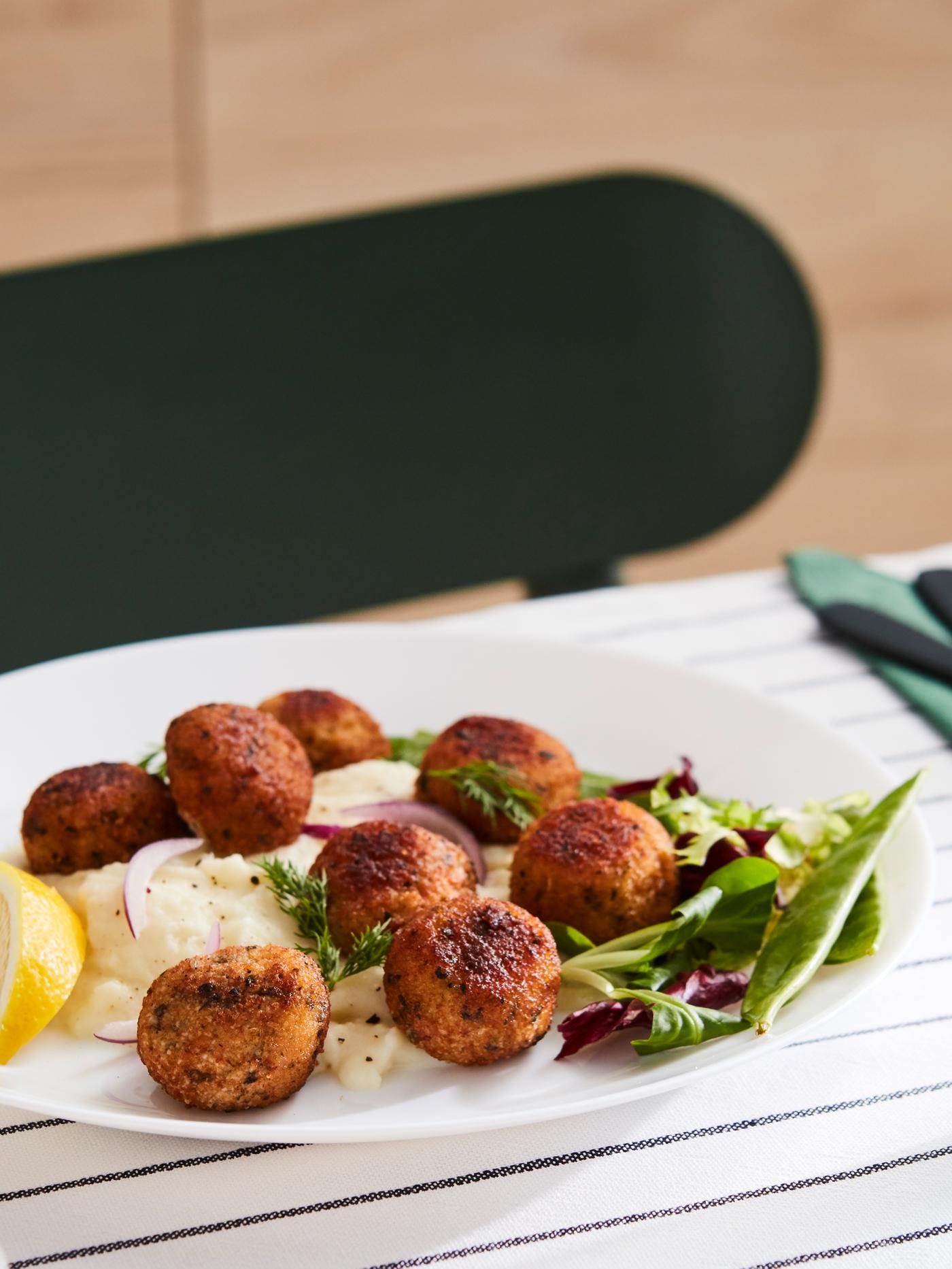 Albóndigas de salmón y bacalao en un plato blanco con puré de papas y ensalada, en una mesa vestida con mantel azul y blanco.
