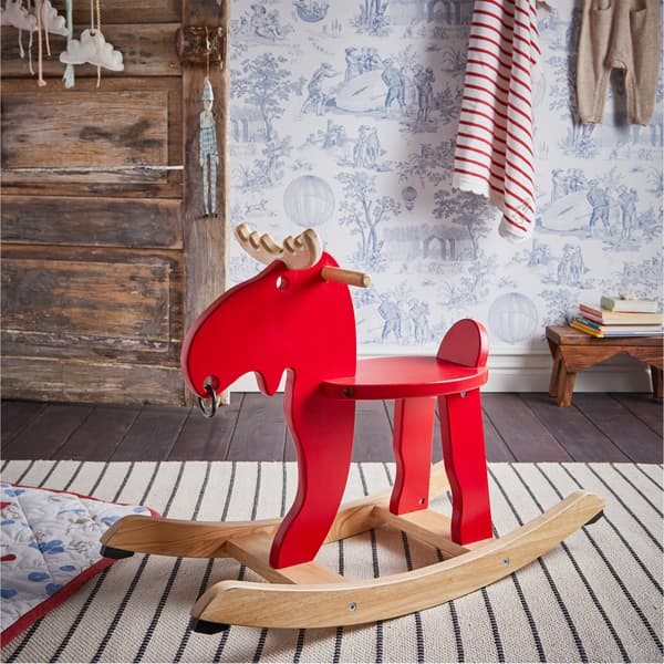 Alce a dondolo EKORRE in rosso/legno dell’albero della gomma su un tappeto bianco a righe nere, accanto a una coperta trapuntata in bianco/rosso/blu – IKEA