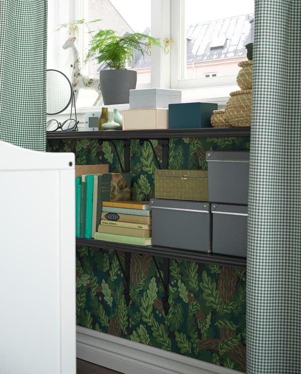 Alcune mensole nere sono fissate alla parete sotto la finestra e utilizzate come comodino. Sulle mensole sono appoggiati libri, scatole e oggetti decorativi - IKEA