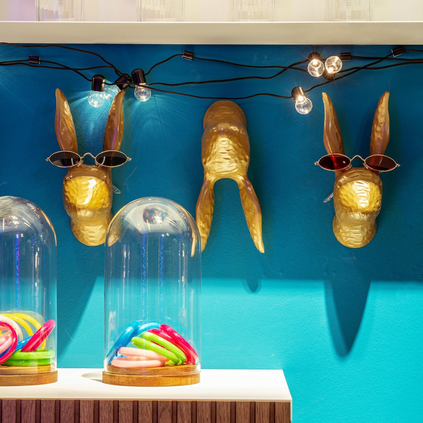 Alcuni ganci oro a forma di coniglio appesi ad una parete azzurra utilizzati come portaocchiali. Davanti, c'è una mensola con campane di vetro BEGÅVNING che contengono elastici per capelli di tanti colori.