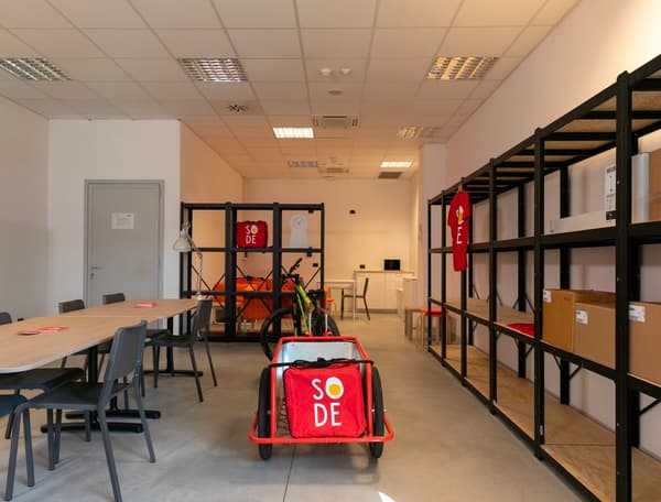 Alcuni scaffali con scatole, un carrello, un tavolo con sedie del So.De – Social Delivery.