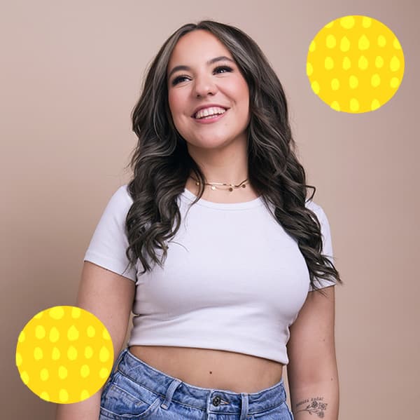 Alisha vor eienr beigen Wand. Sie trägt ein weißes T-Shirt und lächelt. 