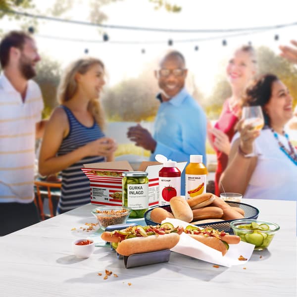 Alle Komponenten des HotDog-Party-Pakets stehen auf einem Tisch, im Hintergrund sind Personen zu sehen, die fröhlich auf einer Gartenparty sind.