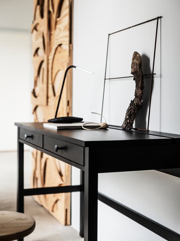 Along a wall in a white room stands a black HEMNES desk and a big, artful wall decoration in untreated wood.