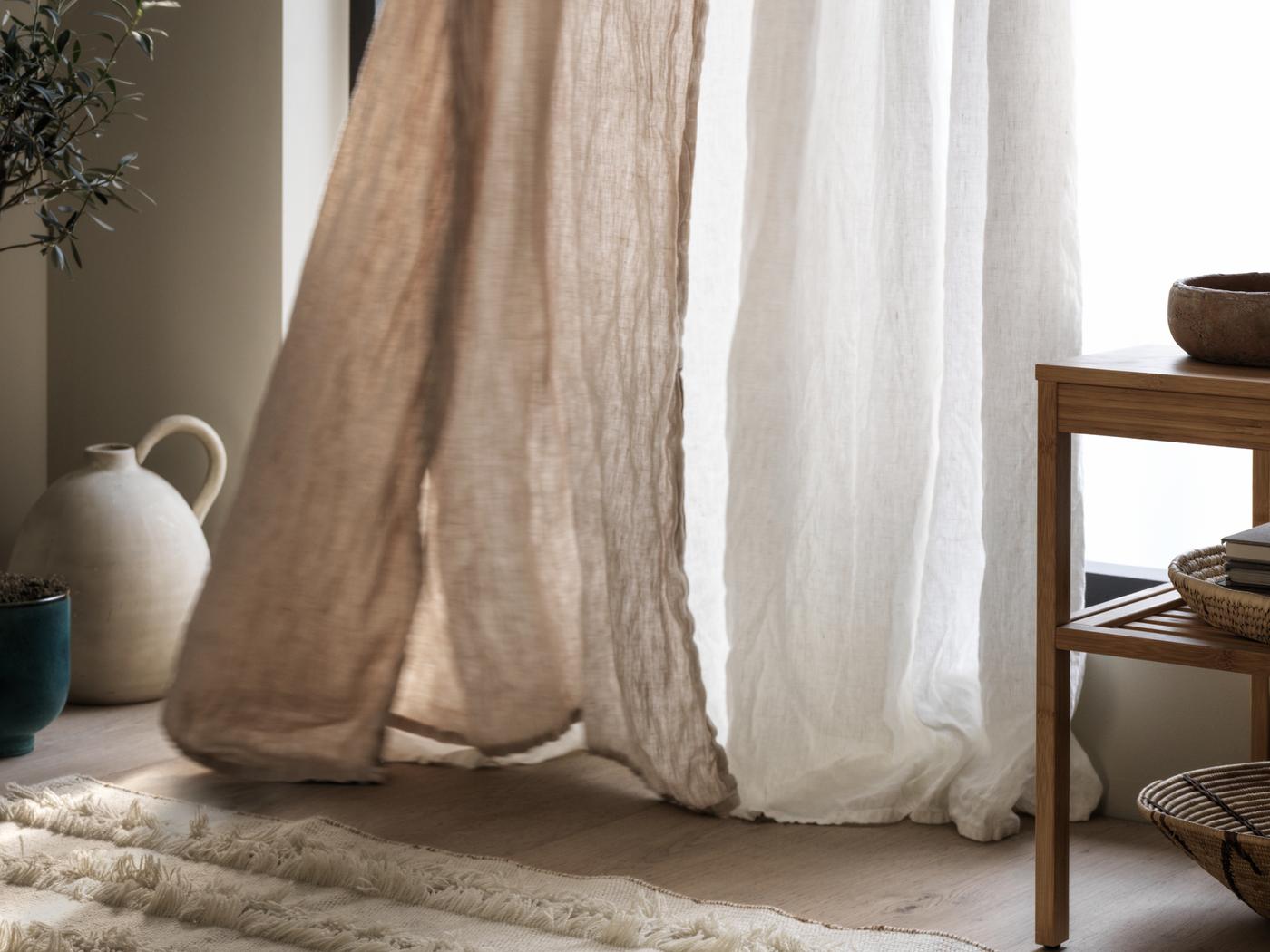 الطرف السفلي من ستارة باللون البيج وستارة SILVERLÖNN شفافة بيضاء تلامس الأرض الخشبية وانتفاخ الهواء أمام النافذة.
