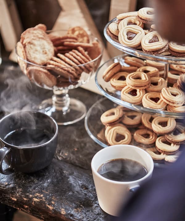 Alzata a tre piani piena di biscotti a forma di cuore, due tazze di caffè caldo e una ciotola con biscotti rotondi.