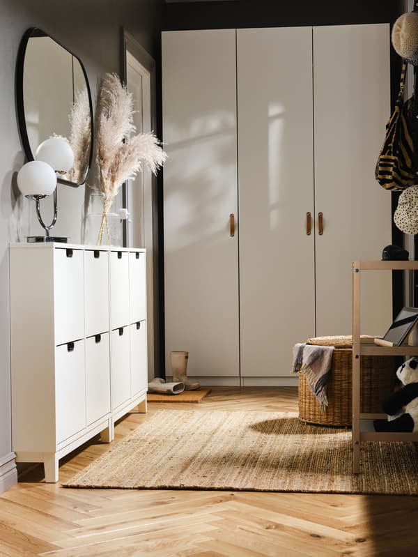 Am Ende eines Vorzimmers ist ein weißer PAX/FORSAND Kleiderschrank mit zwei weißen STÄLL Schuhschränken an einer Seite zu sehen. Über diesen hängt ein Spiegel.