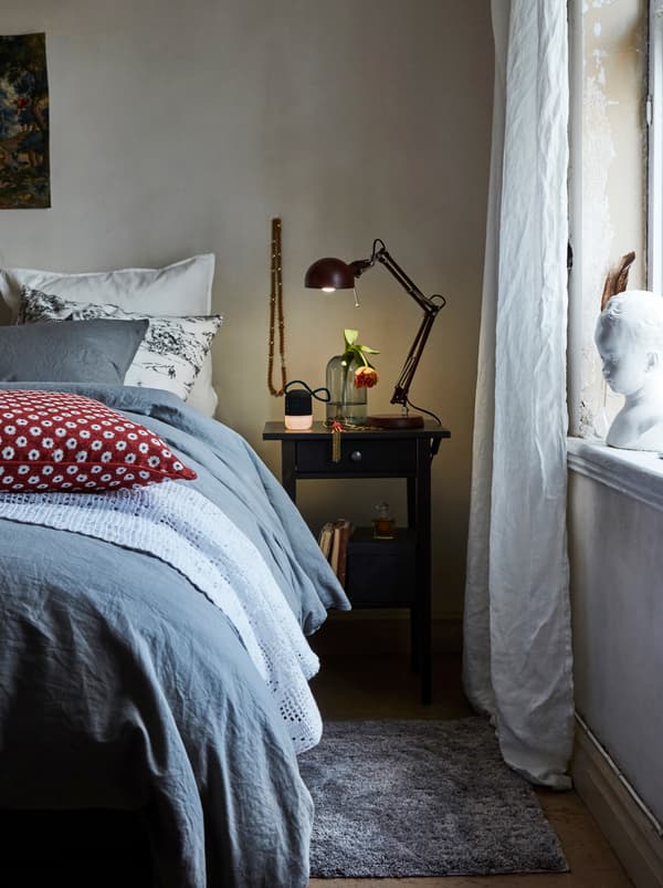 Ambal berwarna kelabu dan meja sisi katil terletak di sebelah katil yang mempunyai bantal beraneka warna, alas katil berwarna lembut dan duvet berwarna biru di atasnya.