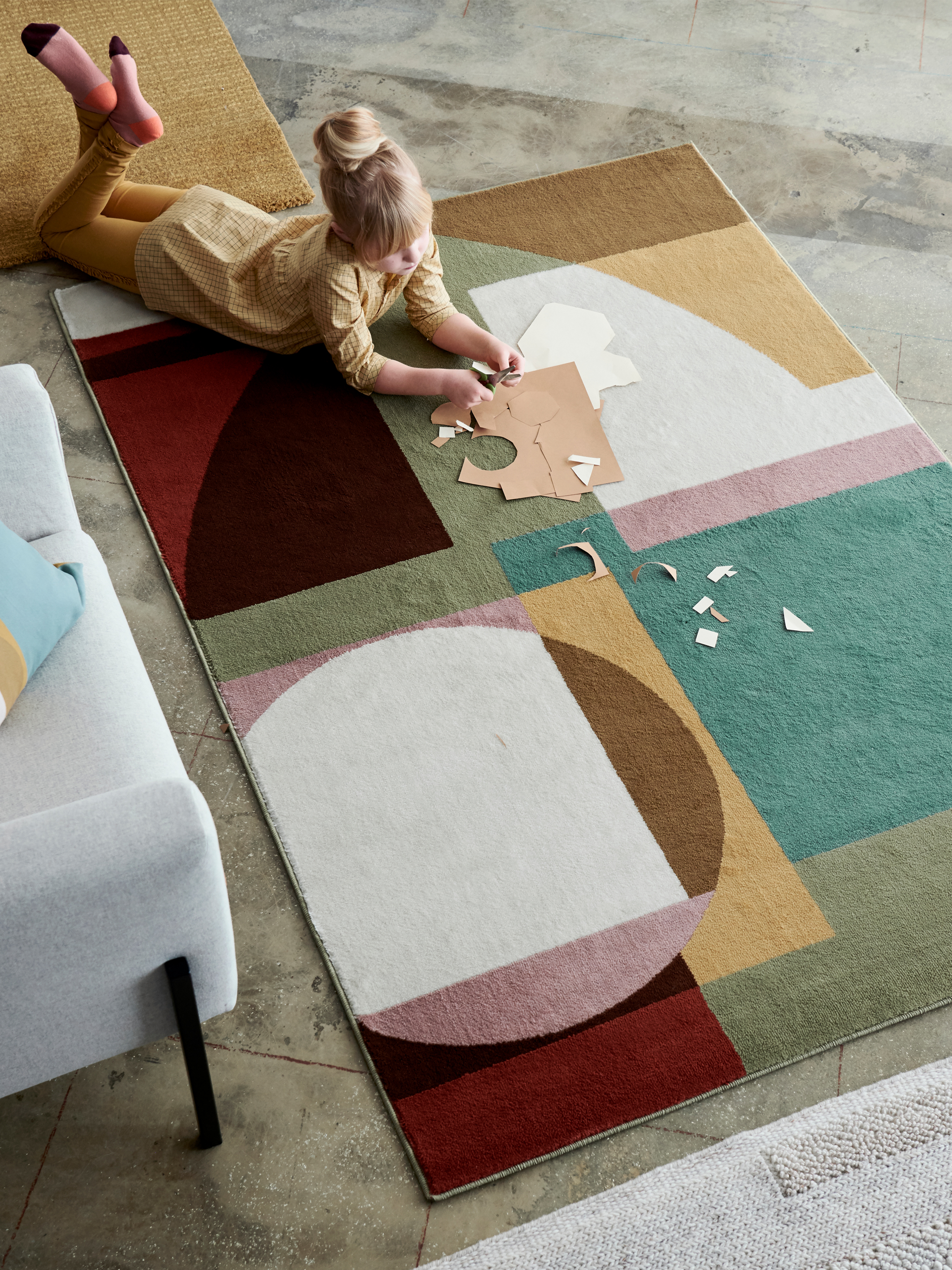 Ambal STENMÄTARE bercorak Bauhaus dalam warna pastel terbentang di tepi sofa dengan seorang budak perempuan di atas ambal tersebut membuat kerja seni dan kraf tangan.