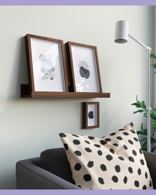 Ambiente de sala de estar con una repisa angosta de madera en la pared que sostiene dos cuadros enmarcados; debajo cuelga un cuadro pequeño. En primer plano, parte de un sillón gris con un cojín beige con puntos negros.