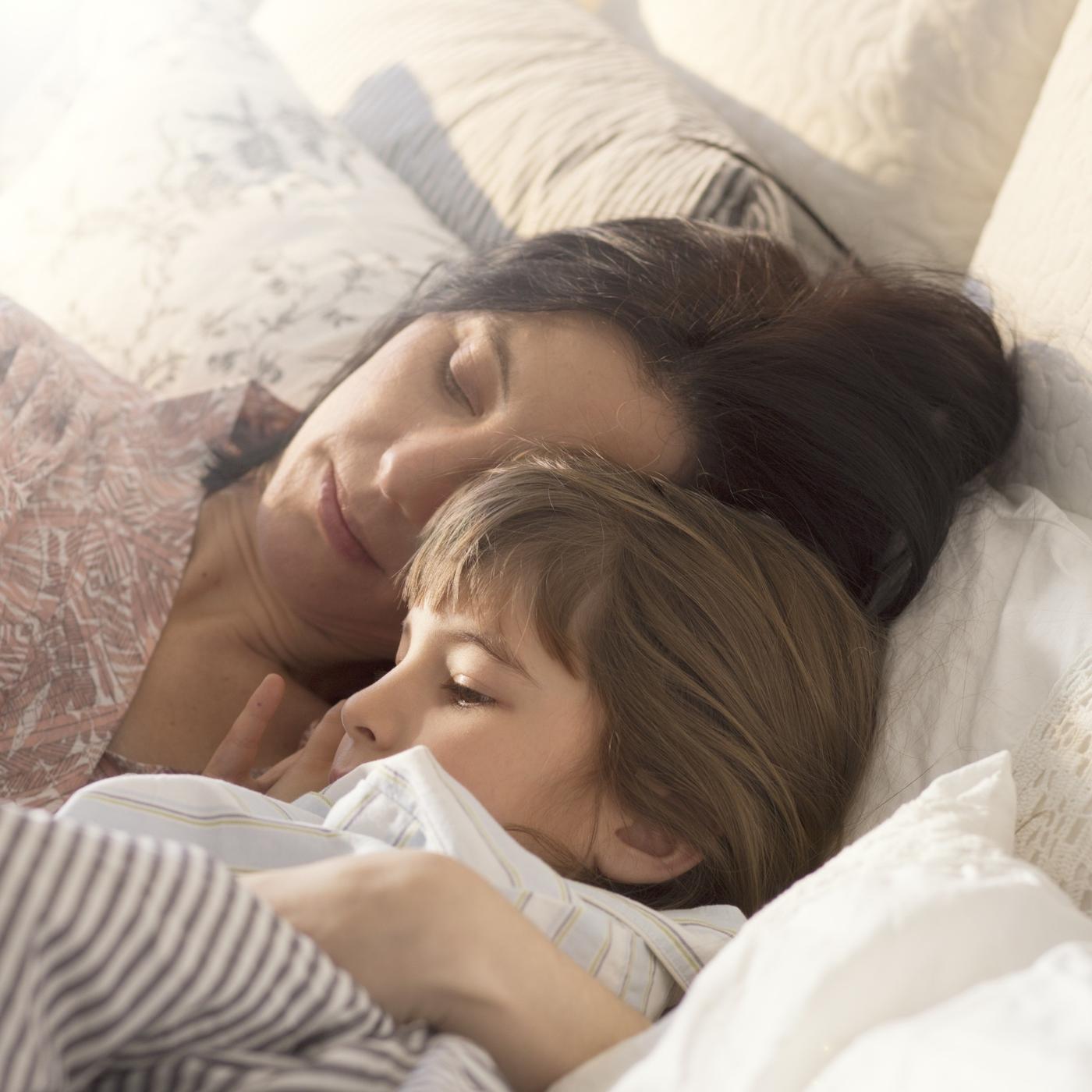 An adult and a child lying in bed together, cuddled under white bedding, both asleep with their heads close.