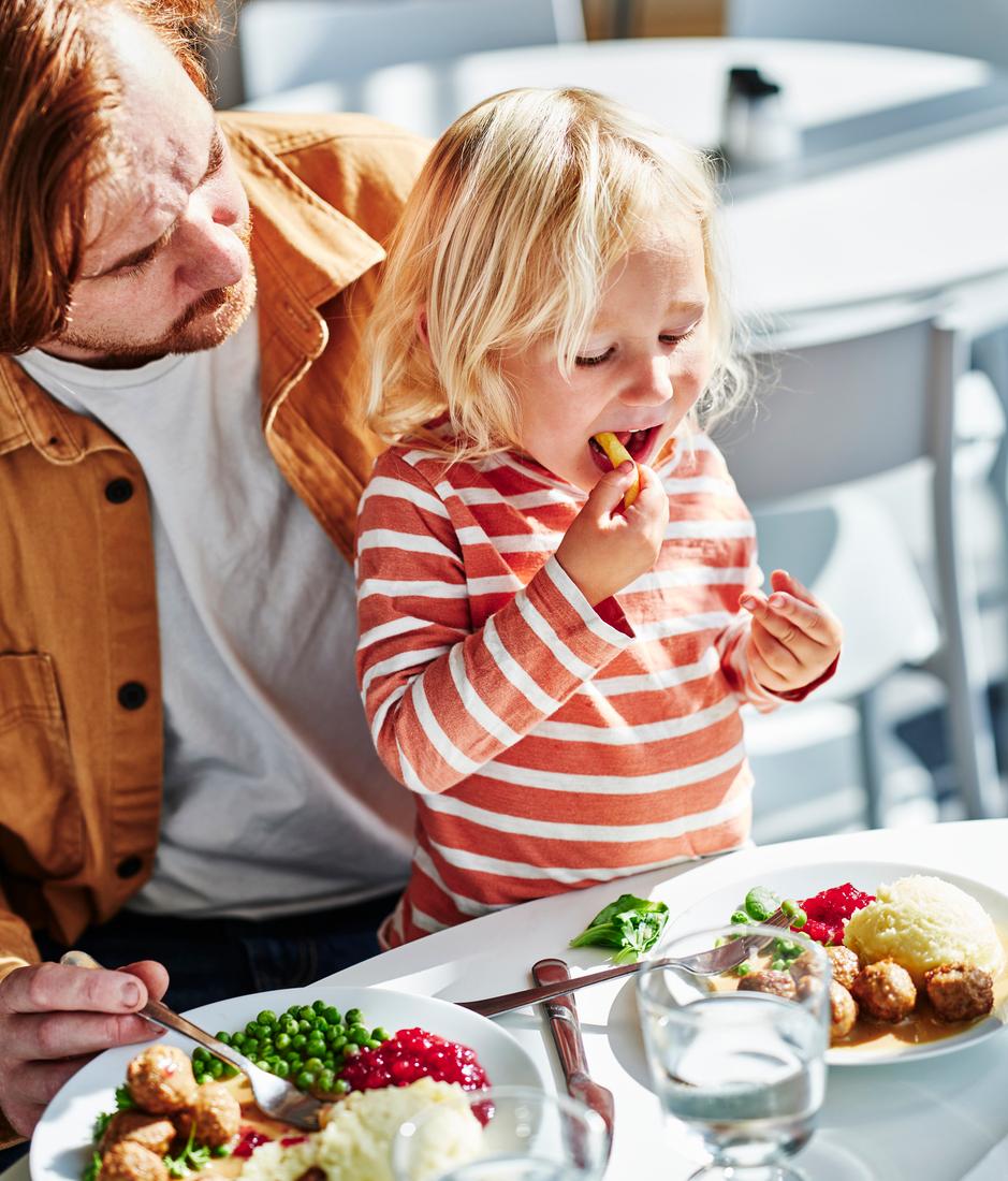 An adult and a young child at a table, with the child eating while plates of meatballs, mashed potatoes, peas, and lingonberries are served in front of them.