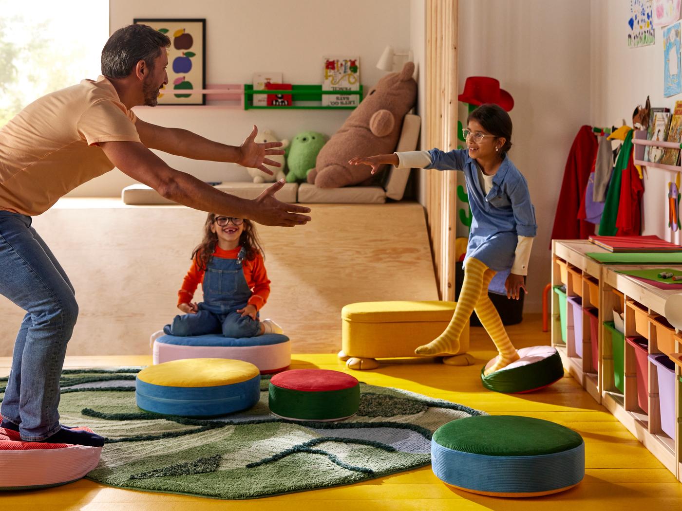 An adult and two children play on colorful round cushions in a playroom with GREJSIMOJS toys, art, and a green carpet on the floor.