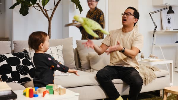 An adult sat on a light beige sofa tosses a green soft toy to a child who stands nearby with outstretched arms.