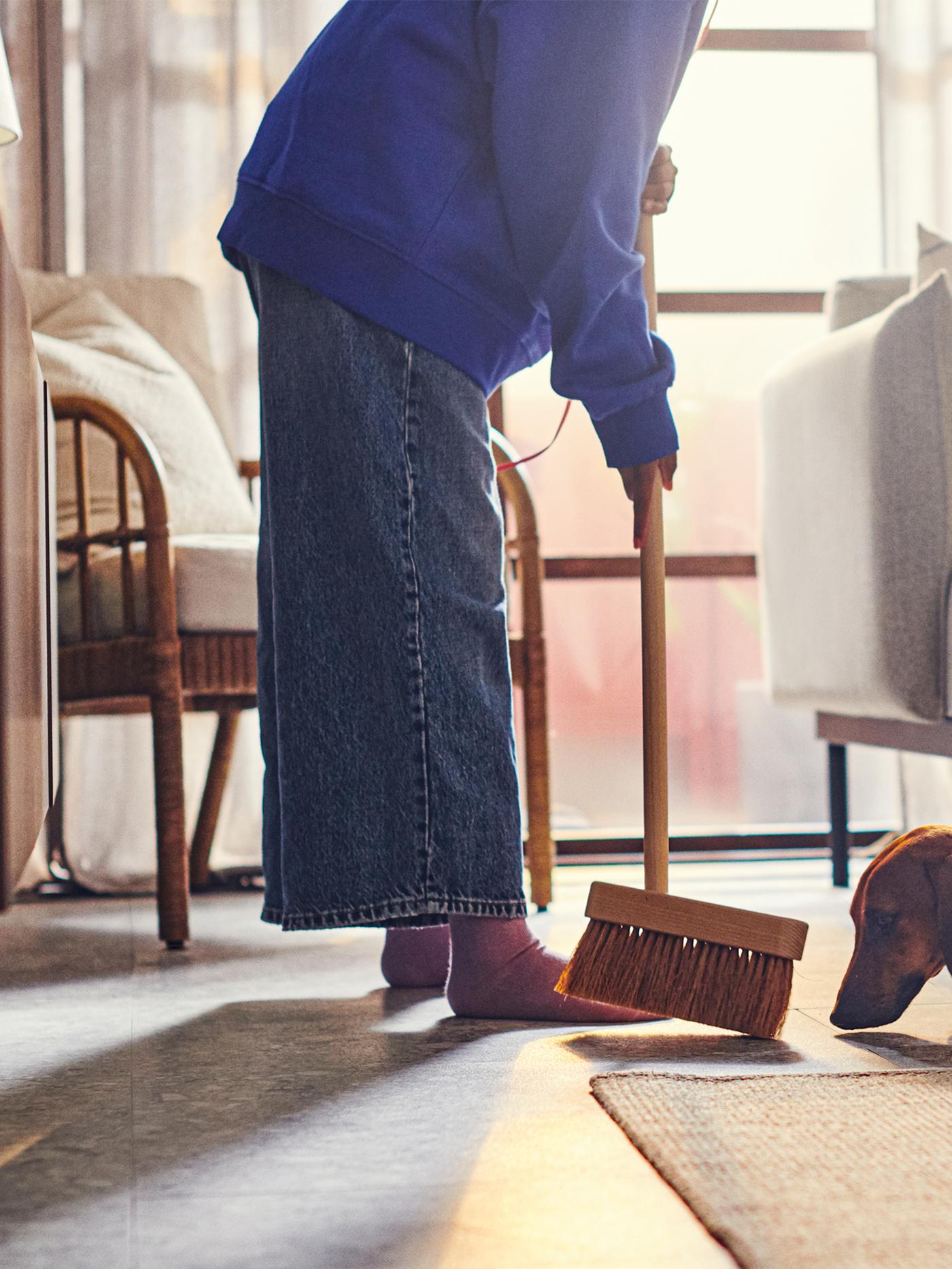 An adult sweeps the floor next to an ÄPPLARYD sofa in light grey and a VODSKOV jute rug. A dog sniffs the broom.