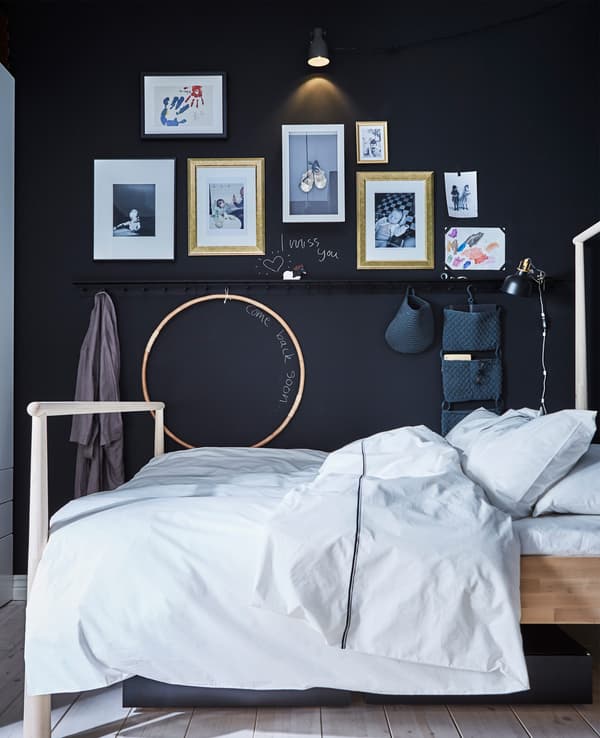 An adult’s bed in against a wall covered in children’s artwork and wall rails with hanging organisers 
