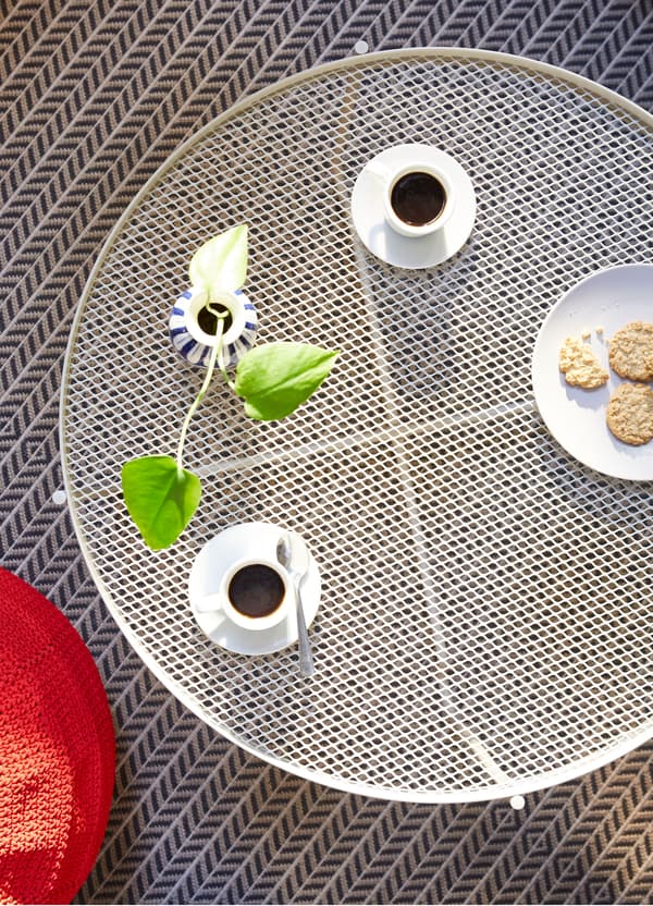 An aerial view of a beige IKEA KROKHOLMEN outdoor coffee table, set with coffee cups, a plant and a plate of cookies.