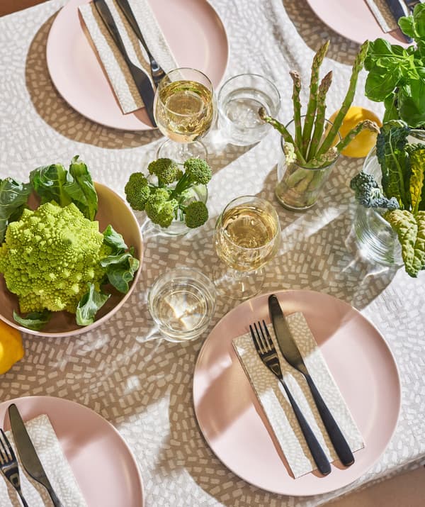 An aerial view of an outside dining table set with pink plates, cutlery, plants and vegetables presented in bowls.