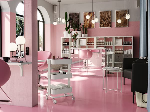 An airy, pink hair salon with white KALLAX shelving behind a mirrored reception desk. A RÅSKOG trolley is by a workstation.