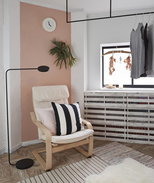An armchair and floor lamp in the corner of a room with a pink feature wall and clothes hanging on a rail across the window.
