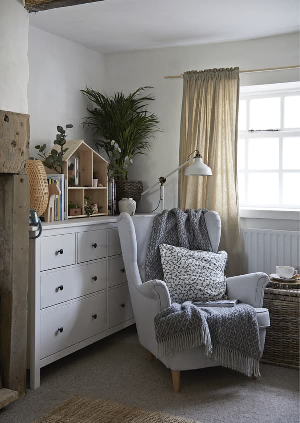 An armchair with throw, cushion and lamp next to a large white chest of drawers.