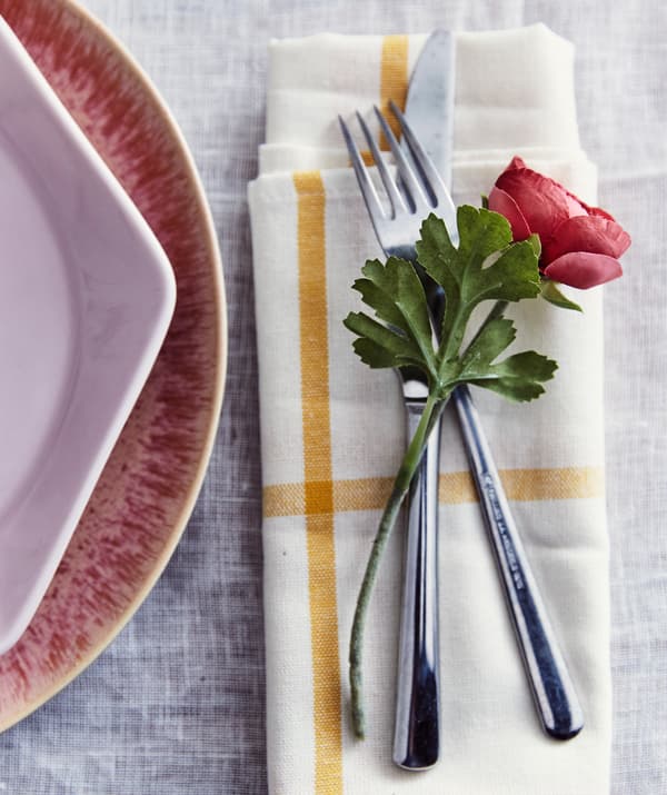 An artifical flower resting on a knife and fork on a yellow and white patterned napkin, next to pink plates.