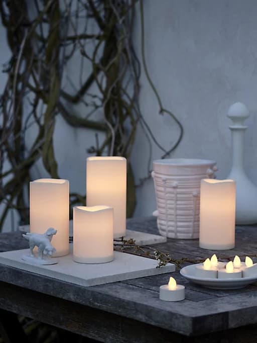 An assortment of LED candles gathered together outside on a wooden table, against a white wall.